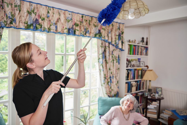 Female Home Help Cleaning House As She Dusts And Talks To Senior Woman